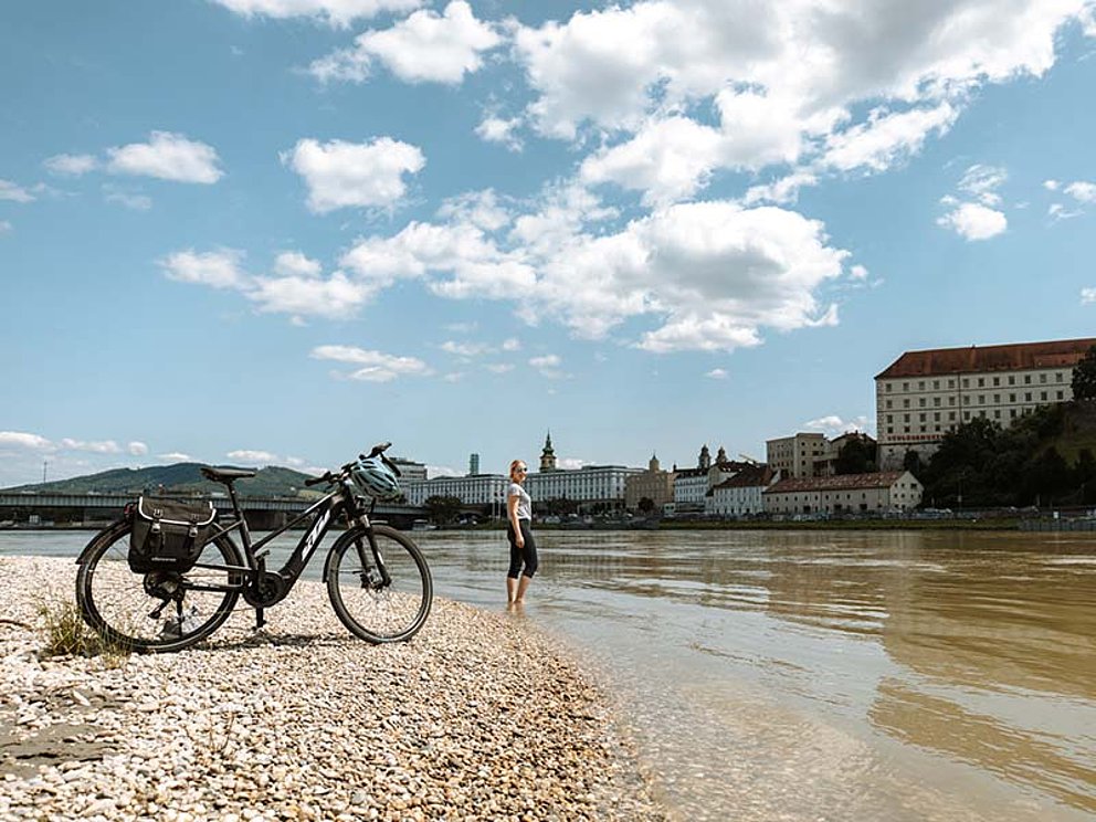 Radfahrerin, die bis zu den Knöcheln in der Donau steht. Im Vordergrund ein E-Bike, im Hintergrund die Stadt Linz