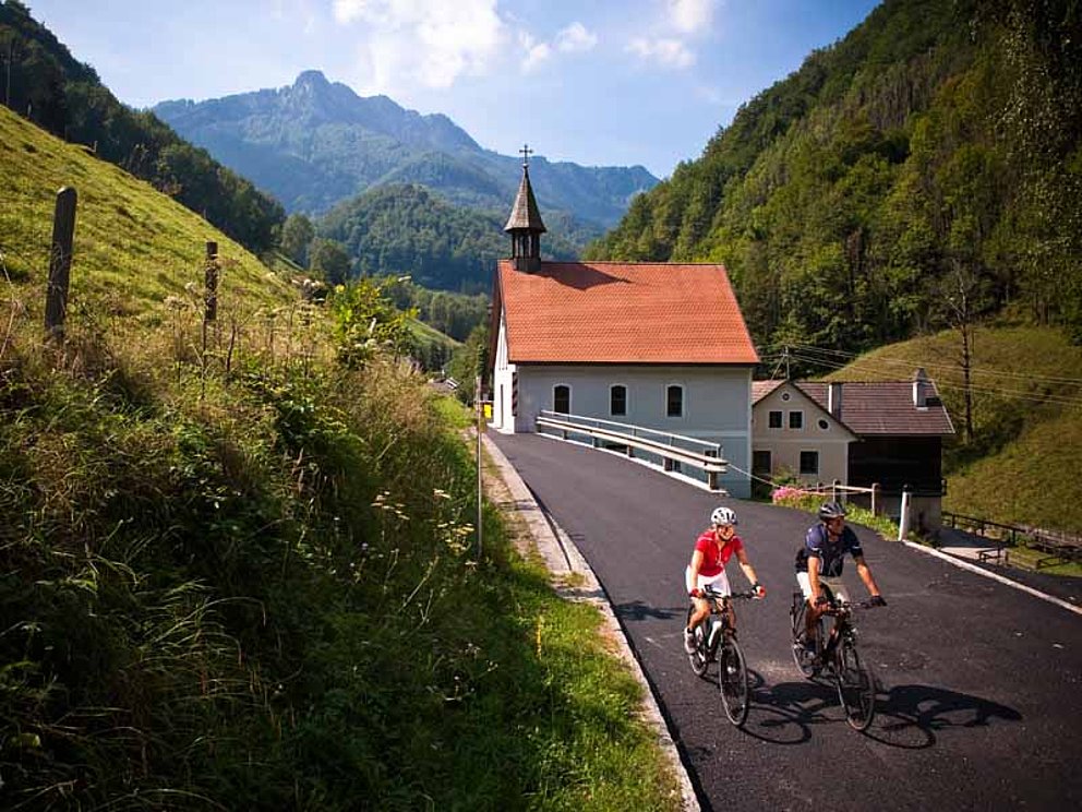 zwei Radfahrer auf einer einsamen Straße nahe der Enns. Am Straßenrand steht eine kleine Kirche.