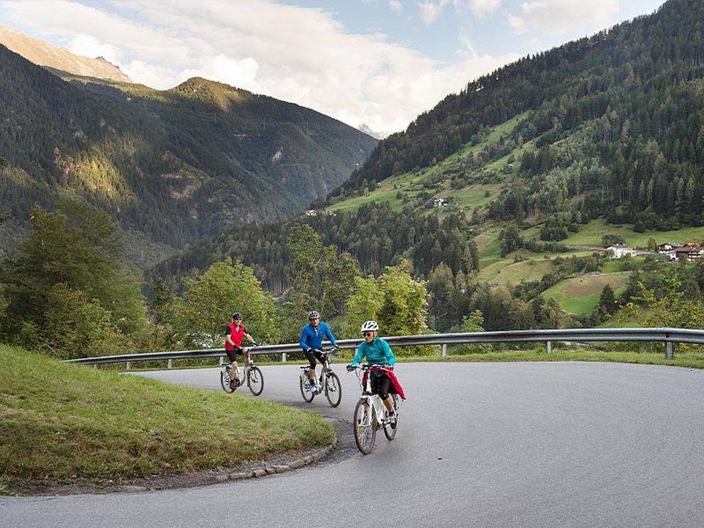 3 Radfahrer radeln bergauf am asphaltierten Radweg, im Hintergrund Bergpanorama