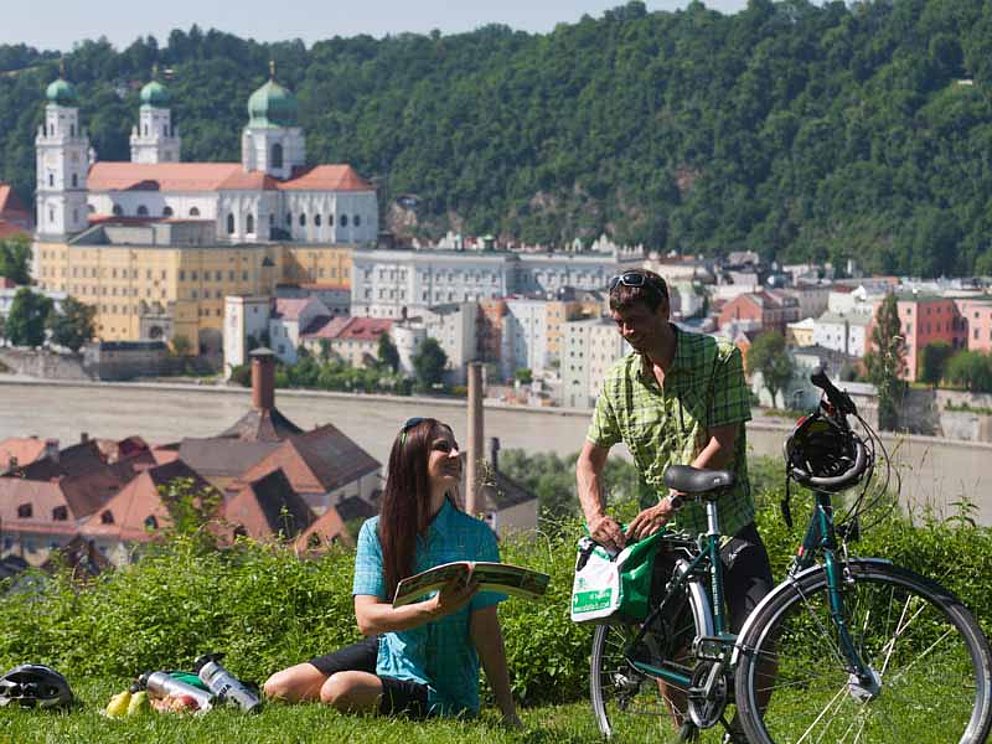 Ein junges Paar bereitet ein Picknick vor. Sie sitzt im Gras und er entpackt die Satteltasche am Fahrrad. Im Hintergrund sind die Türme des Domes in Passau zu sehen.