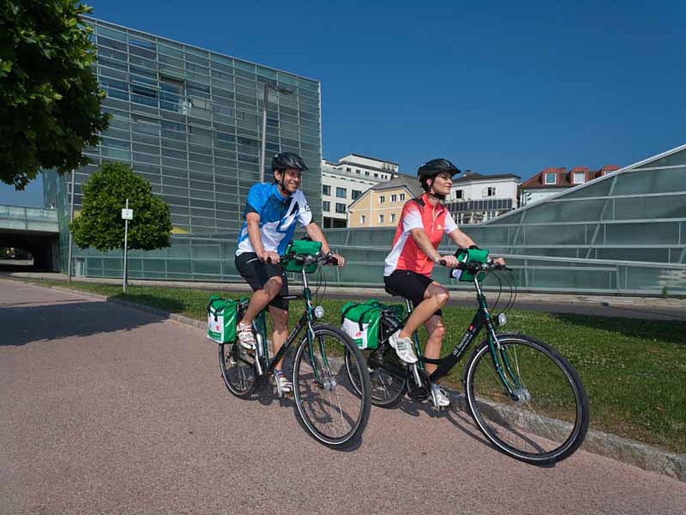 Zwei Radfahrer auf dem Donauradweg in Linz. Im Hintergrund die Glasfassade des Ars Electronica Centers.