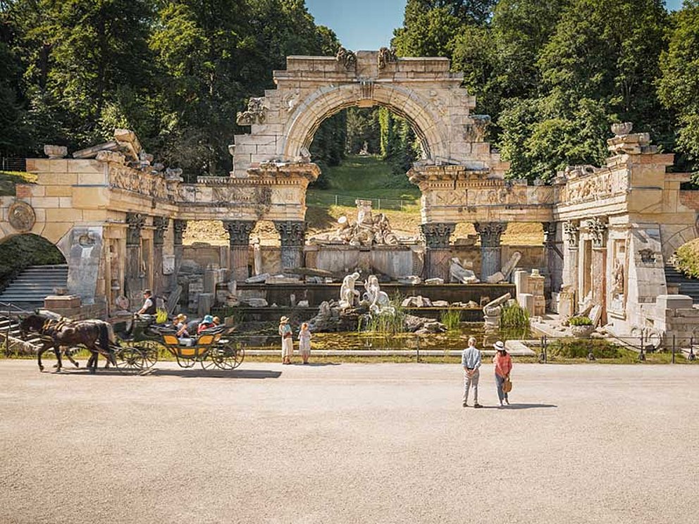 Die Römische Ruine mit Säulen, Bögen und Statuen im Schlosspark Schönbrunn. Davor Besucher und ein Fiaker