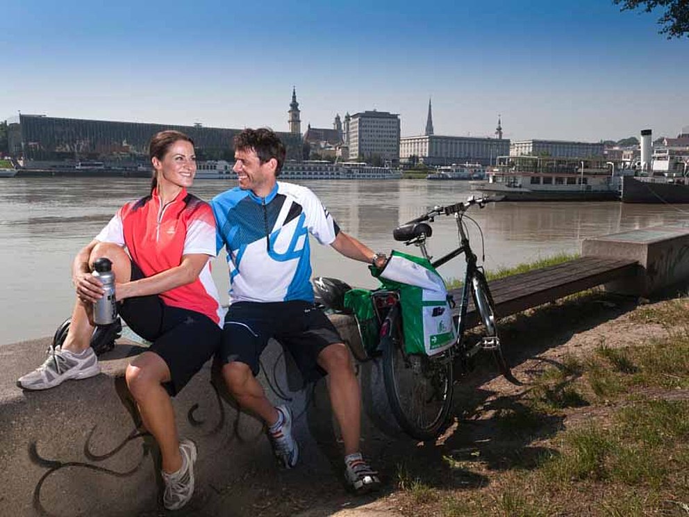 Ein junges Paar hält eine Rast auf einer Mauer am Donauufer in Linz. Rechts steht ein Fahrrad. Vom anderen Ufer grüßt die Stadt.