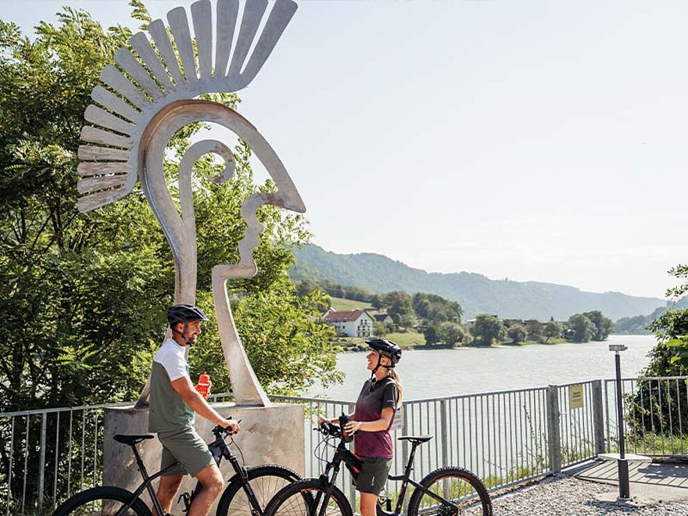 Zwei Radfahrer vor einer modernen römischen Helm-Skulptur am Römerburgus Oberranna mit Blick auf die Donau und die Hügellandschaft.