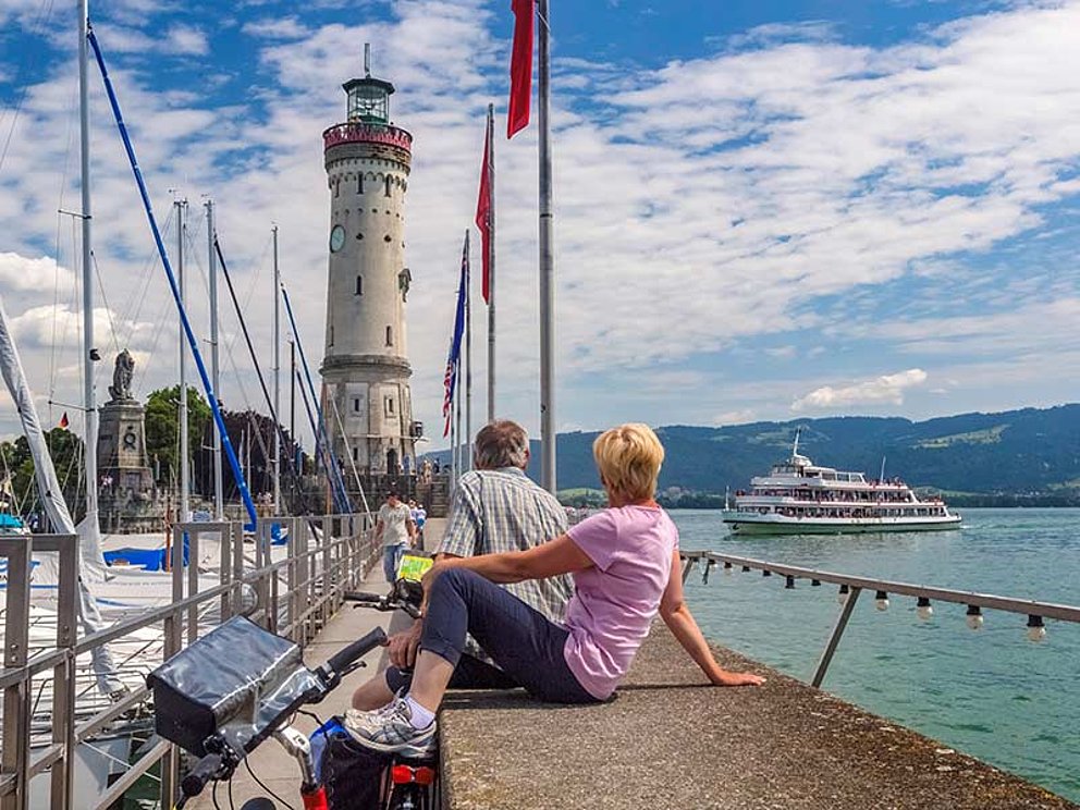 a couple sits at the landing stage in front of the lighthouse, sailing boats nearby
