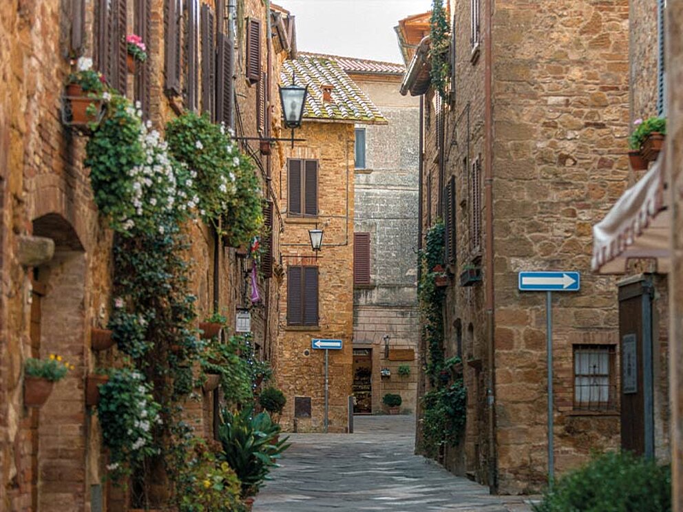 Malerische, schmale Gasse in Pienza, Toskana, mit blühenden Blumen an den historischen Steinmauern.