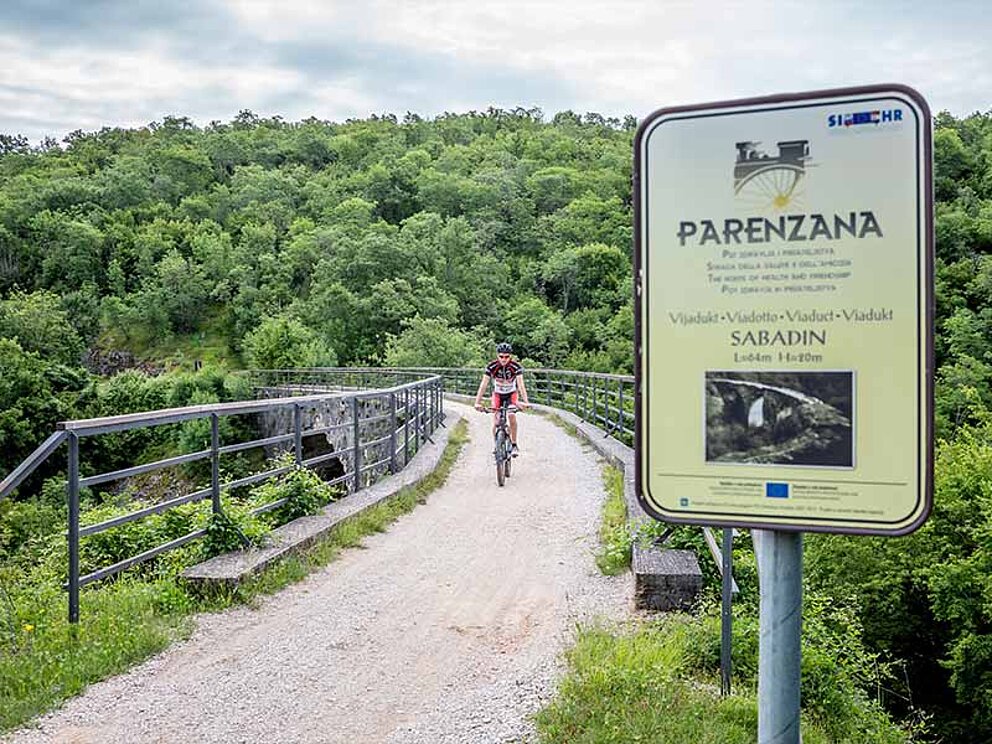Schild der Parenzana, eine Brücke mit Geländer und Biker