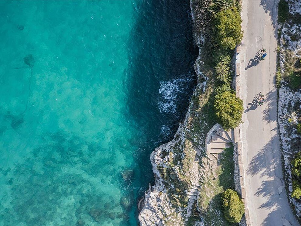 Luftaufnahme von zwei Radfahrern auf einer Küstenstraße in Apulien, Italien, direkt neben kristallklarem türkisfarbenem Meer und hellen Felsklippen.