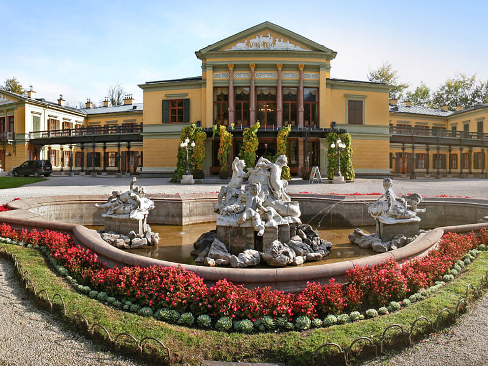 Die Kaiservilla in Bad Ischl. Ein großer Brunnen mit Statuen steht auf dem Vorplatz. blühende Blumenbeete umrahmen den Brunnen.