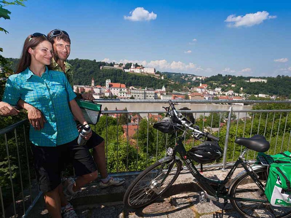 Ein junger Mann hält eine junge Frau im Arm. Sie stehen auf einer Aussichtsplattform hoch über Passau. Daneben steht ein Fahrrad mit Gepäcktasche.