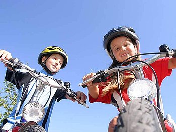 Zwei Kinder mit Helmen und Raddressen gucken über ihre Fahrradlenker in die Kamera. Der Fotograf hat die Aufnahme vom Boden aus nach oben gemacht.