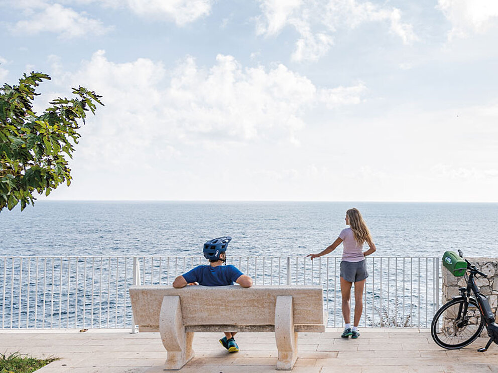Ein Paar blickt von einer weißen Terrasse über das weite blaue Mittelmeer in Apulien, ein Fahrrad steht daneben.