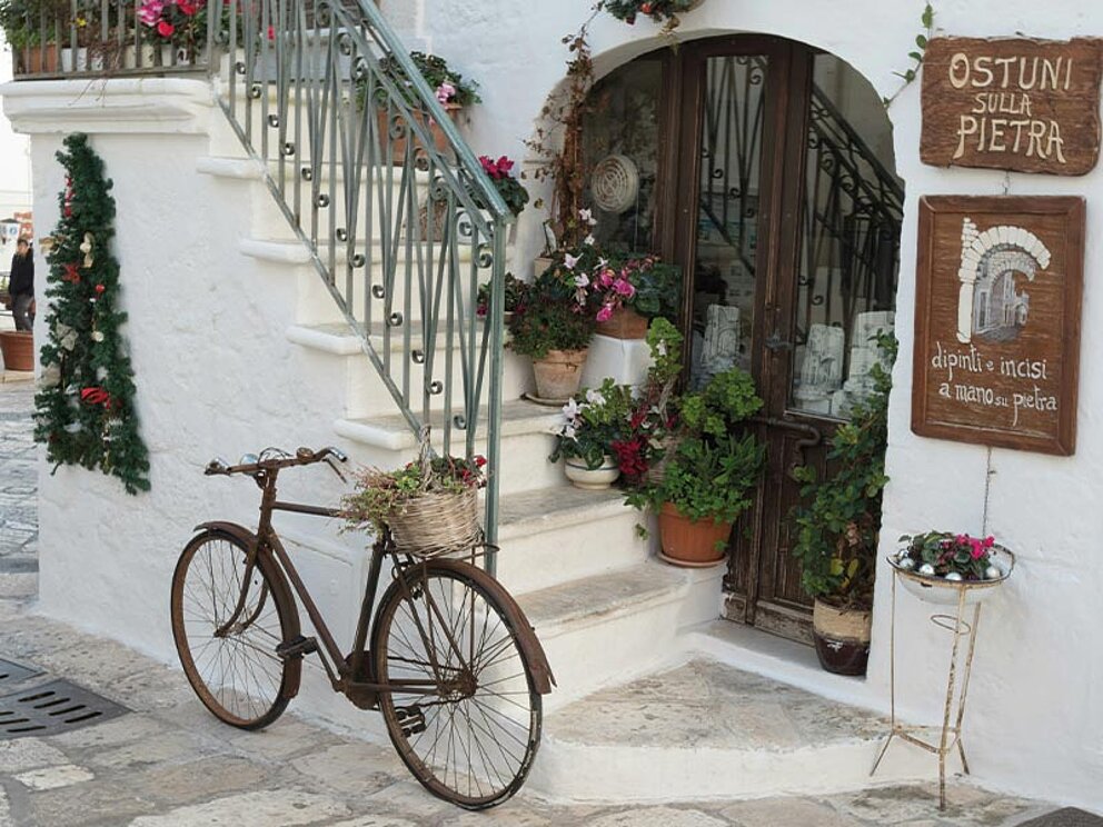 Ein nostalgisches Vintage-Fahrrad vor einem weiß gekalkten Gebäude in Ostuni, Apulien, mit blühenden Blumen und einem Schild „Ostuni sulla Pietra“.
