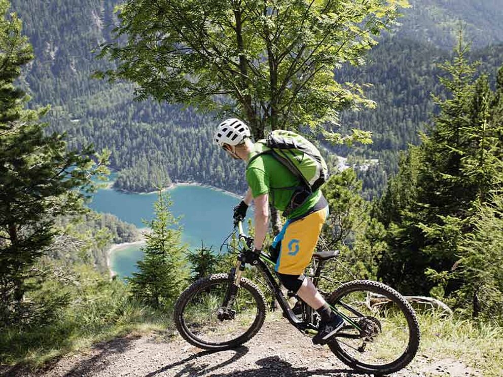 Ein Biker mit Rucksack auf einem Schotterweg, im Hintergrund ein Bergsee