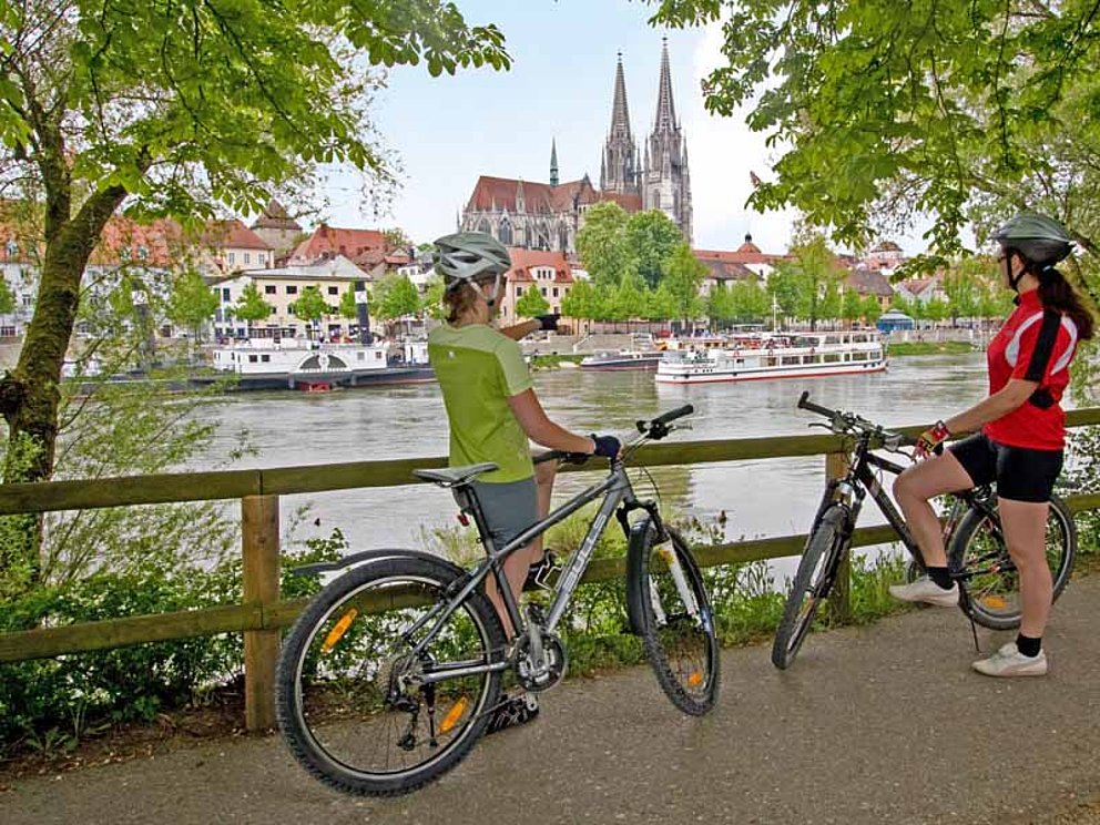 zwei Radfahrer halten am Donauufer in Regensburg. Die Bäume geben den Blick auf die Stadt mit dem markanten Dom am anderen Ufer frei. 