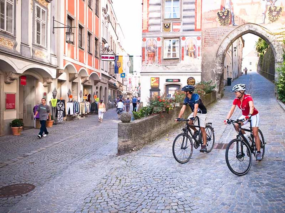 zwei Radfahrer in der Altstadt von Steyr. Die Gassen sind mit Kopfsteinpflaster ausgelegt. Hinter den beiden ist ein Tordurchfahrt. Häuser mit historischen Fassaden dominieren das Stadtbild.