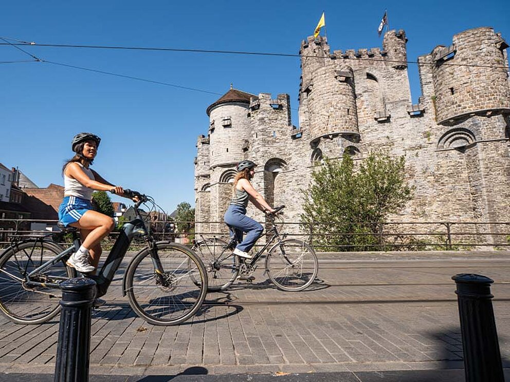 Zwei Radfahrerinnen mit Helmen vor der mittelalterlichen Burg Gravensteen in Gent, Belgien.
