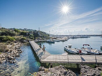 Radfahrer auf einem hölzernen Steg an einer malerischen Küste in Apulien, Italien, mit Booten im glitzernden Wasser und strahlender Sonne.