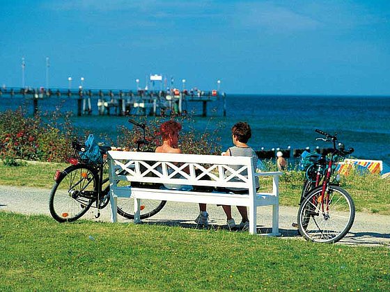 2 Damen sitzen auf einer Bank und blicken auf die Ostsee. 2 Fahrräder stehen daneben