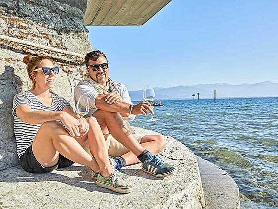 2 Personen sitzen auf einem Stein am Bodenseeufer mit einem Glas Weißwein in der Hand