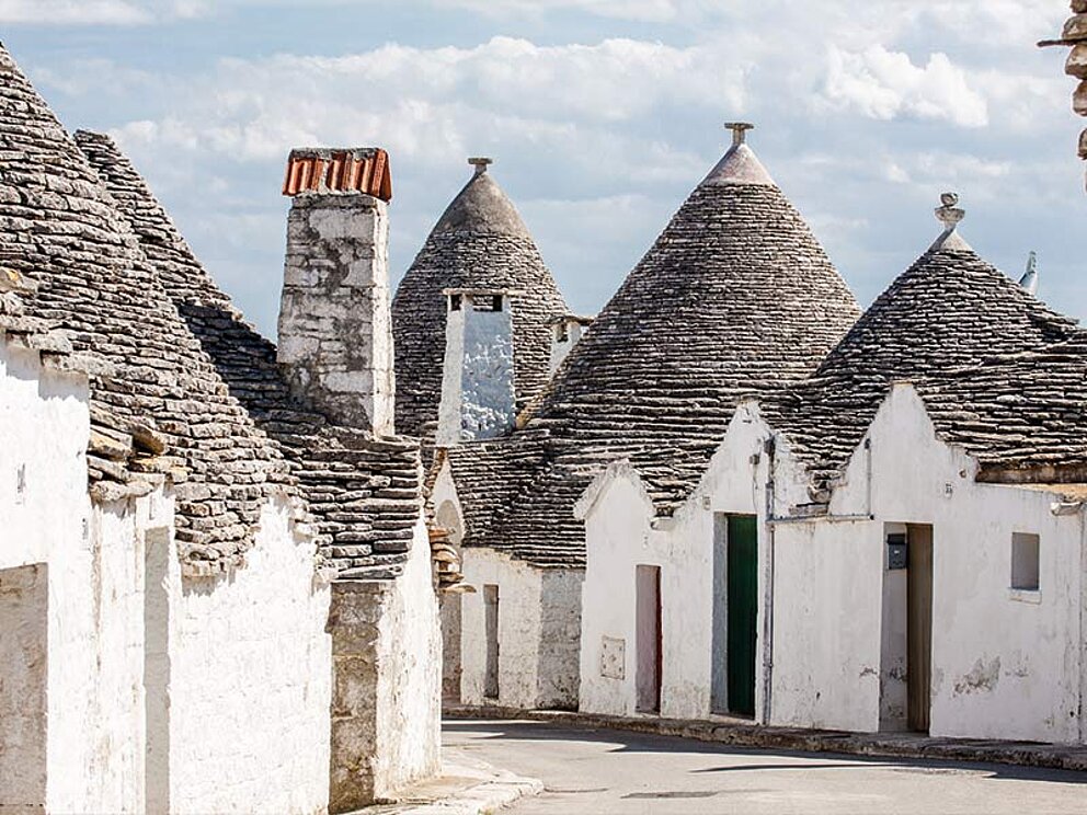 Eine Reihe traditioneller, weiß gekalkter Trulli-Häuser mit den charakteristischen kegelförmigen Steindächern in einem Dorf in Apulien, Italien.