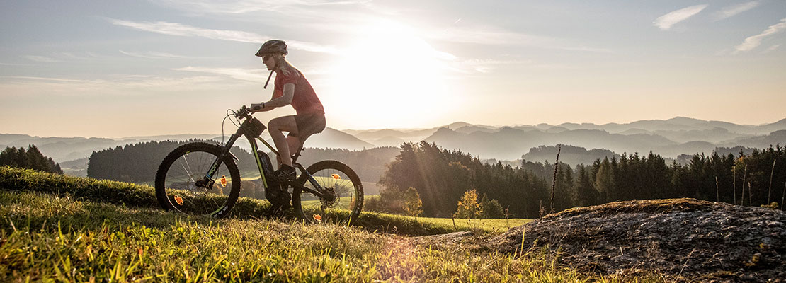 Person auf E-Mountainbike radelt in den Sonnenuntergag mit Panoramablick auf die hügelige Umgebung.
