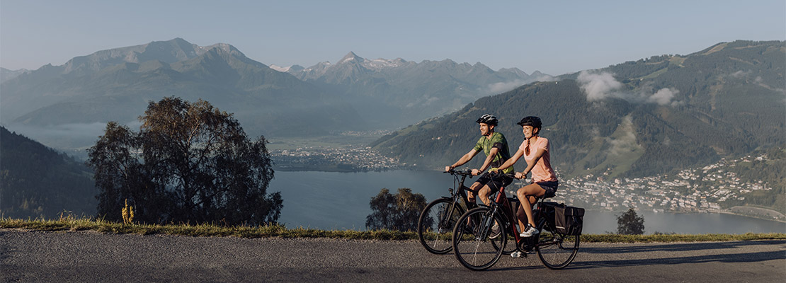 2 Radfahrer am asphaltierten Radweg, im Hintergrund der Zeller See und Berge