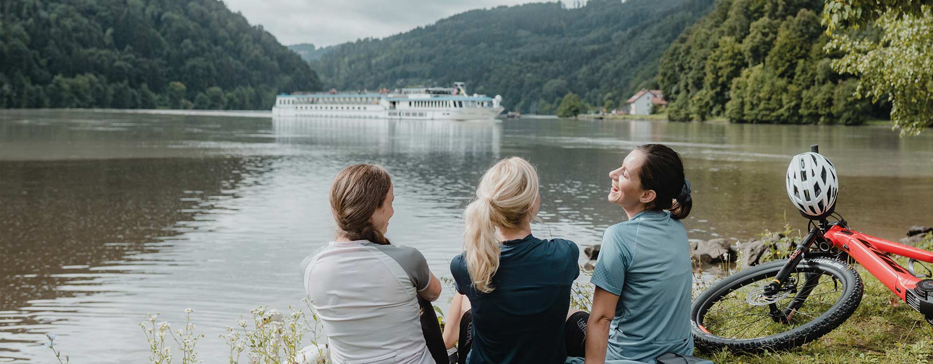 3 Damen sitzen am Donauufer, auf der Donau fährt ein Fluskreuzfahrtschiff