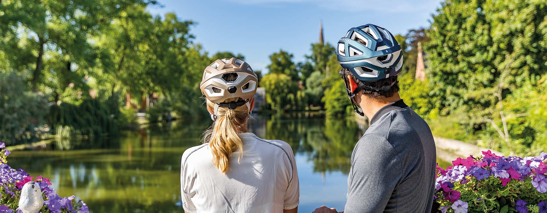 Zwei Radfahrer mit Helmen blicken von einer Brücke aus auf einen ruhigen Kanal in der belgischen Natur.