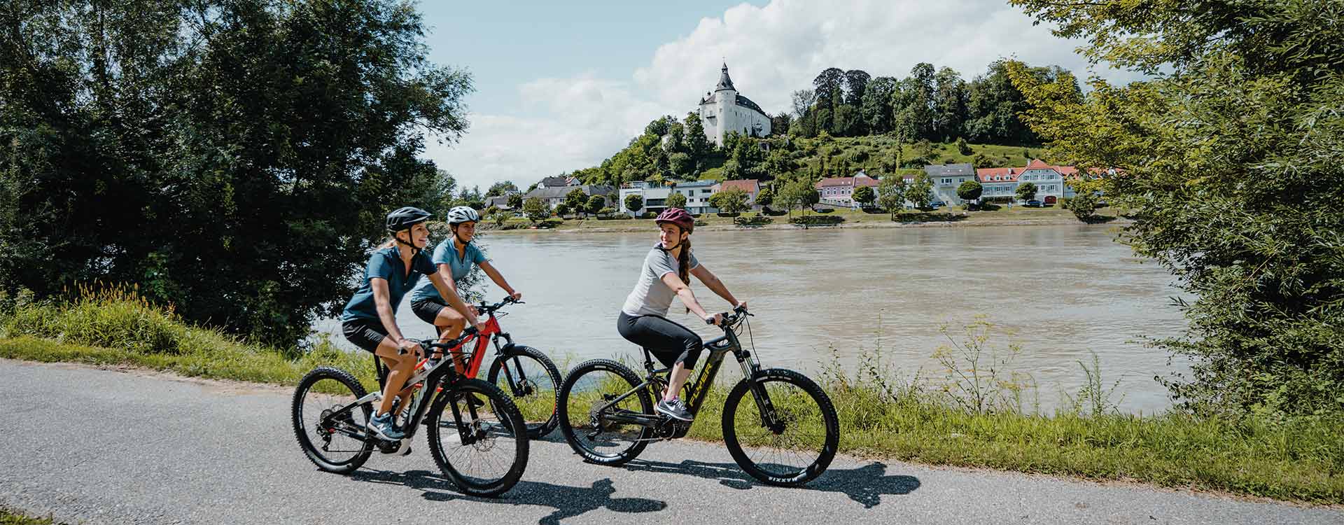 3 Radfahrerinnen am Donauradweg, am anderen Ufer der Ort Ottensheim.